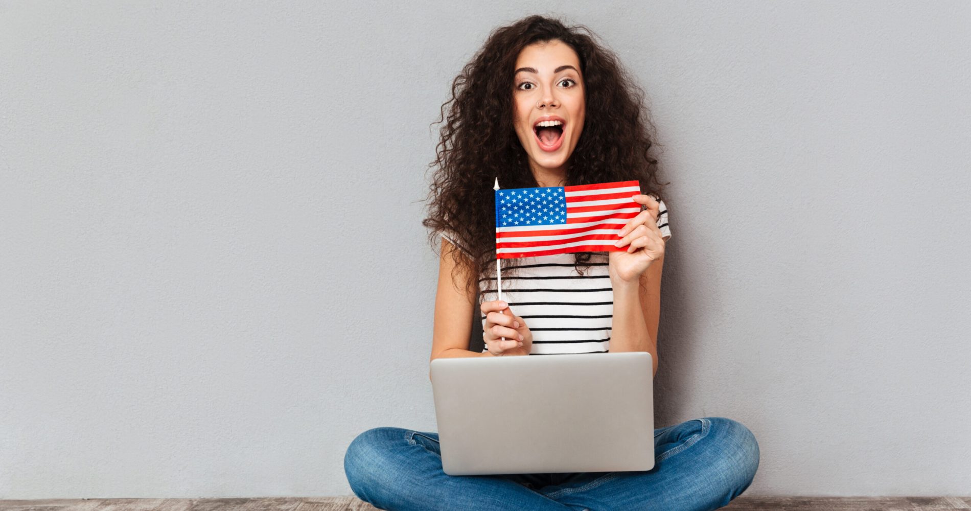 Gorgeous female with beautiful smile sitting in lotus pose with silver computer on legs, demonstrating american flag on camera over grey wall